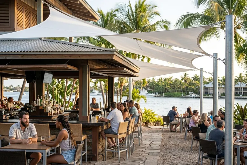 Open-air waterfront bar with white shade sails anchored to the bar roof and steel posts over the seating